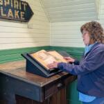 Woman in a blue jacket reads an open old book on a tilted display at a museum setting.