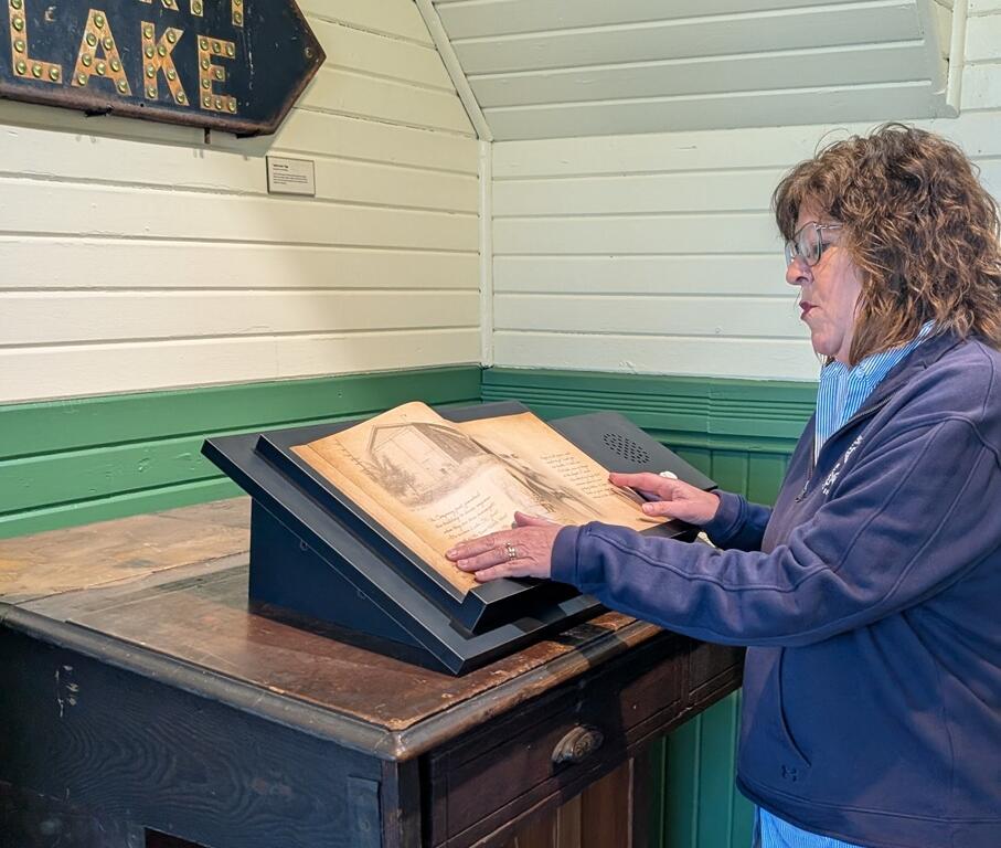 Woman in a blue jacket reads an open old book on a tilted display at a museum setting.