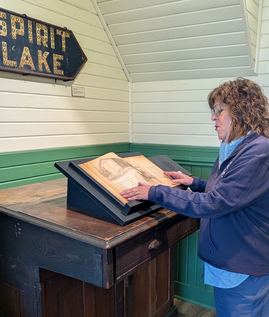 Woman in a blue jacket reads an open old book on a tilted display at a museum setting.