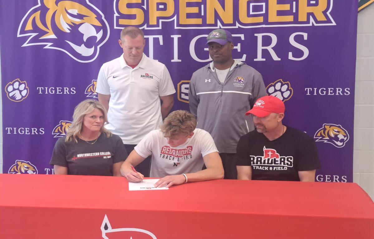 Athlete signs a document at a red table while four people watch, backdrop reads 'Spencer Tigers' with tiger logos.