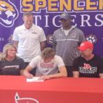 Athlete signs a document at a red table while four people watch, backdrop reads 'Spencer Tigers' with tiger logos.