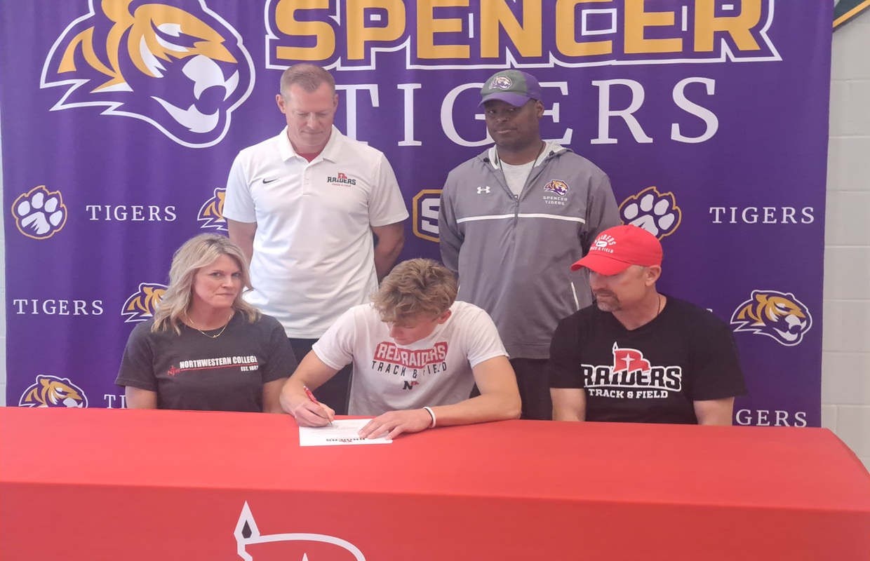 Athlete signs a document at a red table while four people watch, backdrop reads 'Spencer Tigers' with tiger logos.