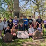 Group of students posing outdoors in front of a memorial monument in a park, some kneeling and others standing, sunny day.