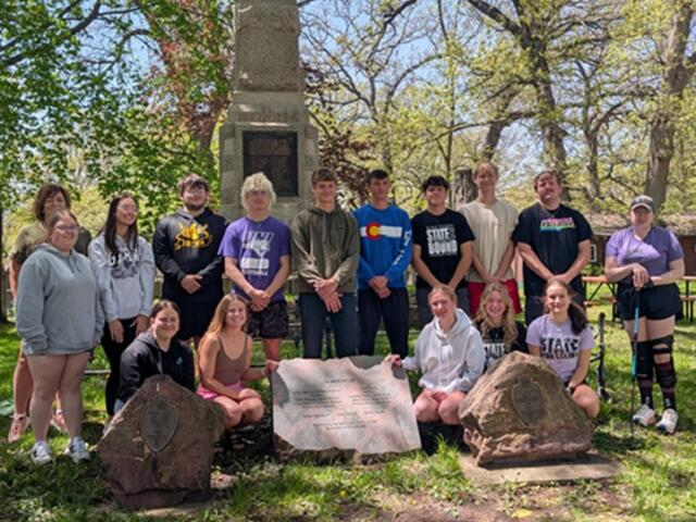 Group of students posing outdoors in front of a memorial monument in a park, some kneeling and others standing, sunny day.