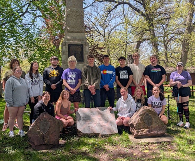 Group of students posing outdoors in front of a memorial monument in a park, some kneeling and others standing, sunny day.