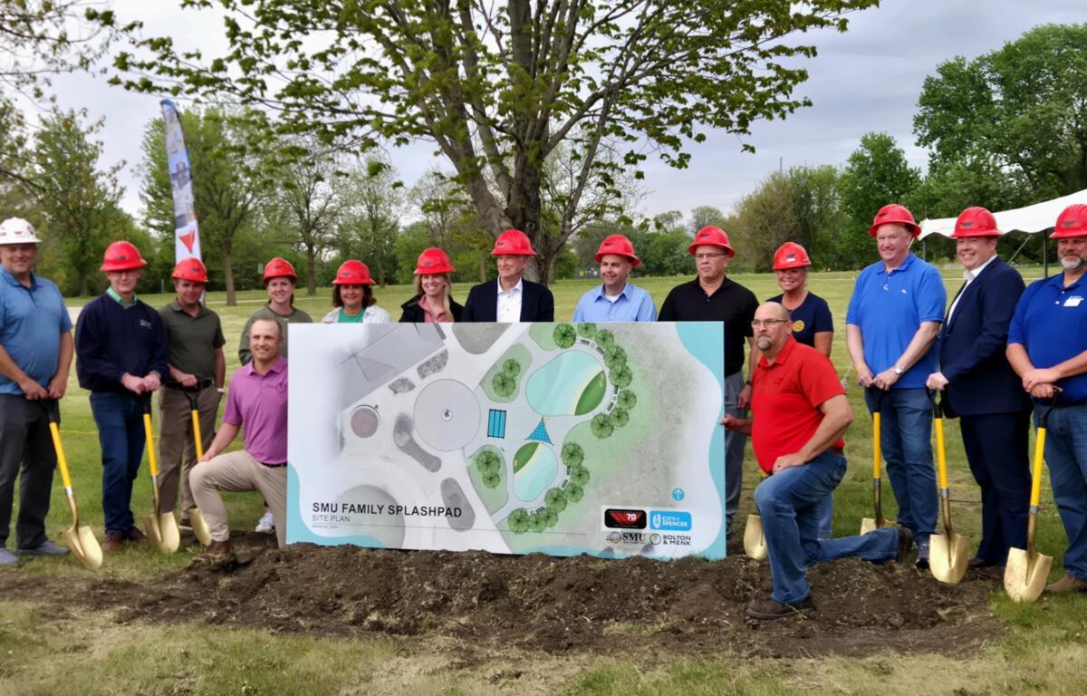 Group of people wearing red hard hats posing outdoors with a large site plan for a splashpad project. Trees and a playground-like site in the background.