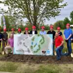 Group of people wearing red hard hats posing outdoors with a large site plan for a splashpad project. Trees and a playground-like site in the background.