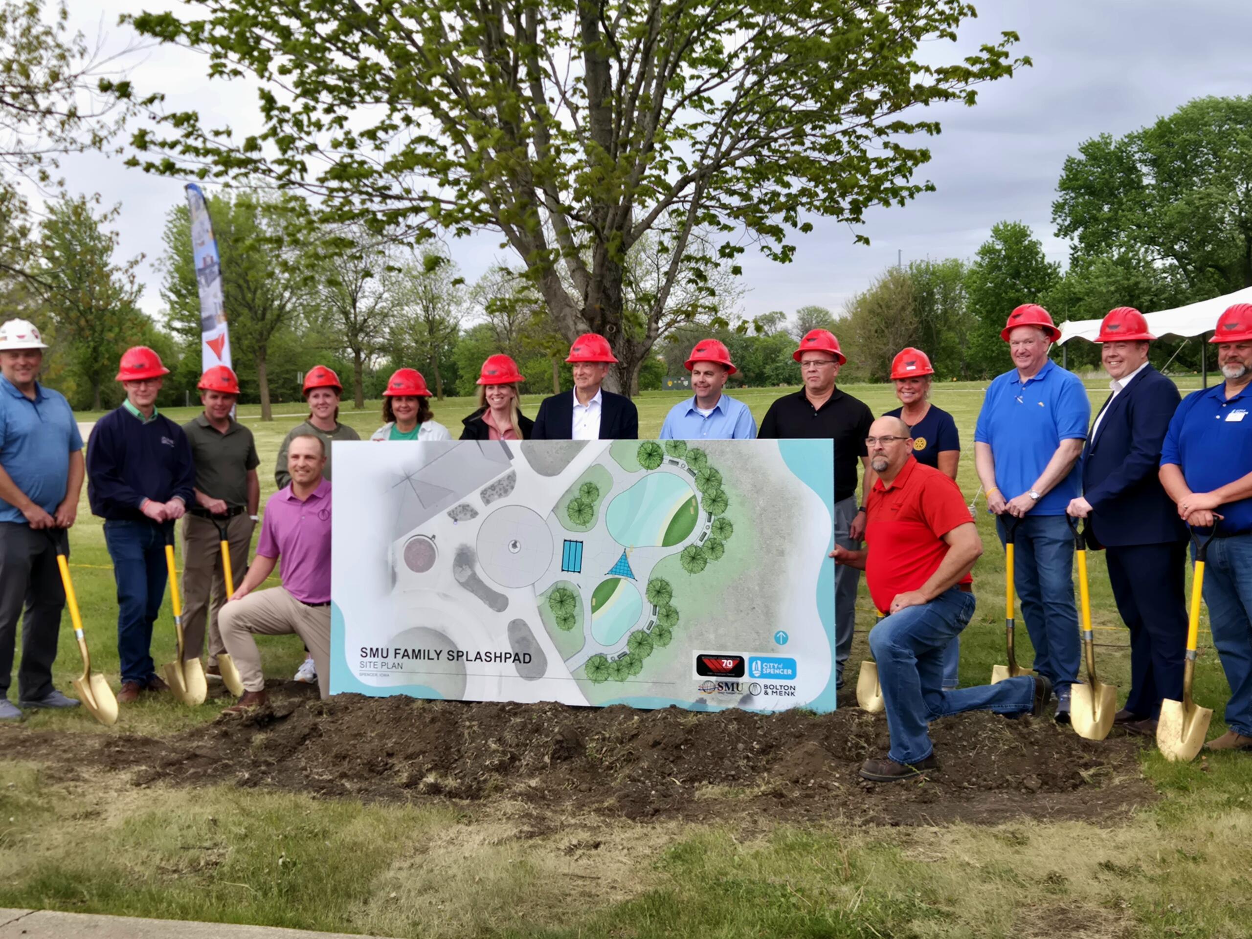Group of people wearing red hard hats posing outdoors with a large site plan for a splashpad project. Trees and a playground-like site in the background.