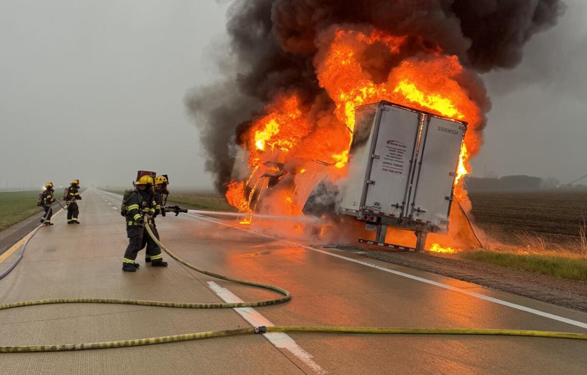 Semi-truck on a highway engulfed in flames with thick black smoke as firefighters spray water from hoses.