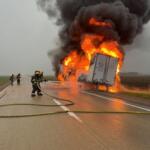 Semi-truck on a highway engulfed in flames with thick black smoke as firefighters spray water from hoses.