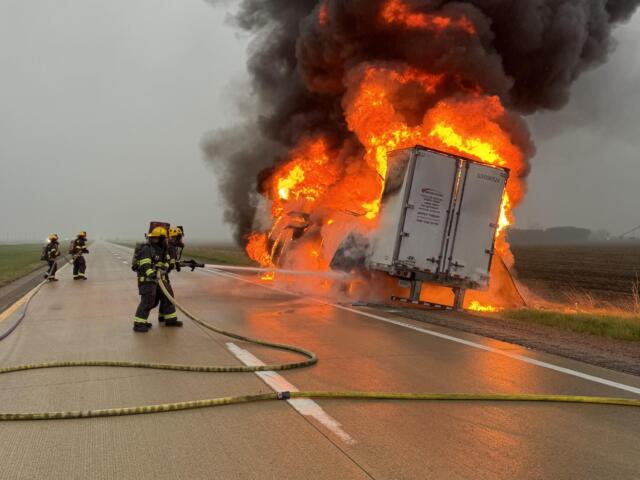 Semi-truck on a highway engulfed in flames with thick black smoke as firefighters spray water from hoses.