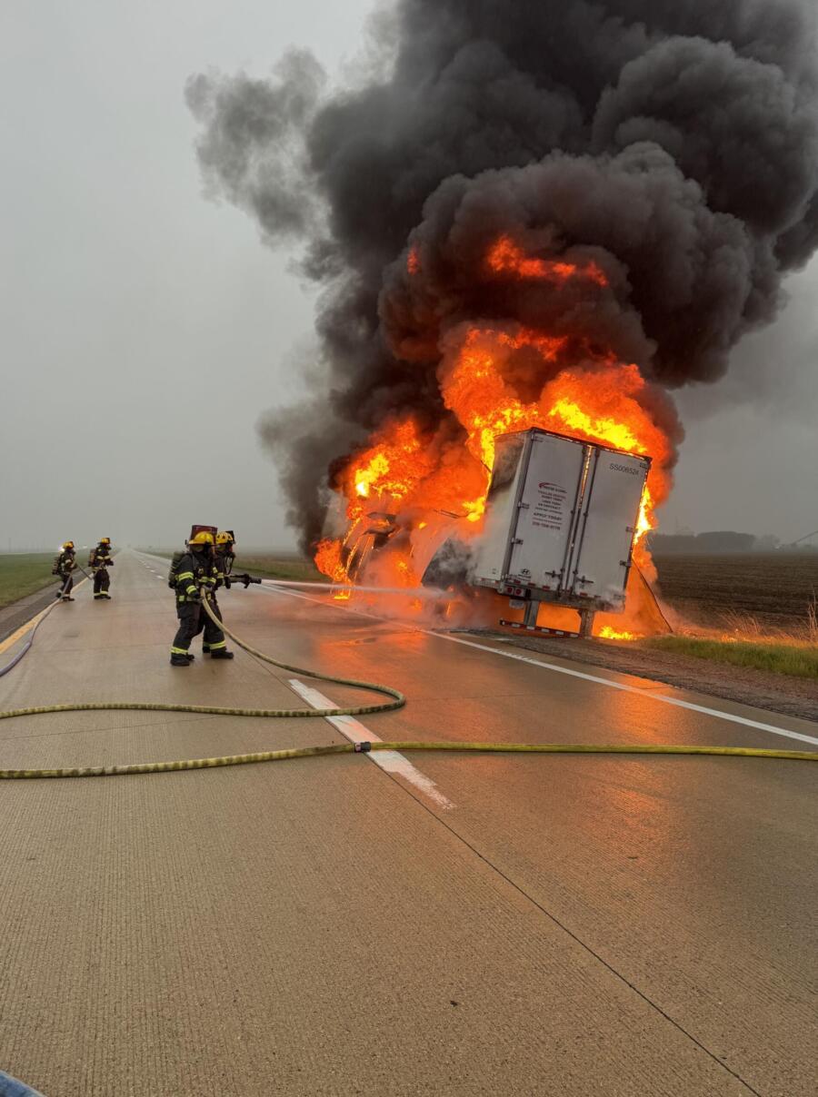 Semi-truck on a highway engulfed in flames with thick black smoke as firefighters spray water from hoses.