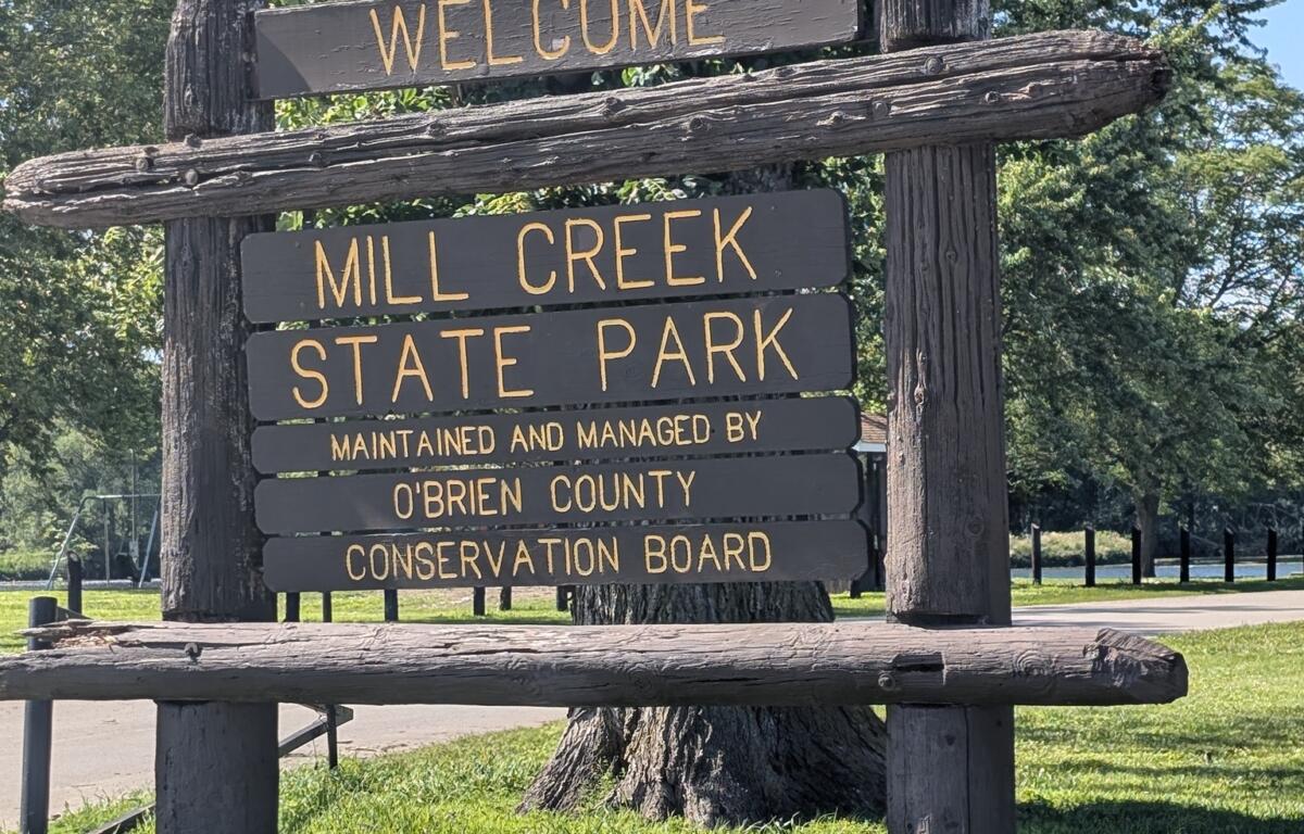Wooden welcome sign for Mill Creek State Park with trees in the background. Maintained by O'Brien County Conservation Board.