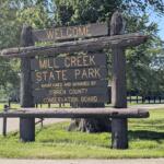Wooden welcome sign for Mill Creek State Park with trees in the background. Maintained by O'Brien County Conservation Board.