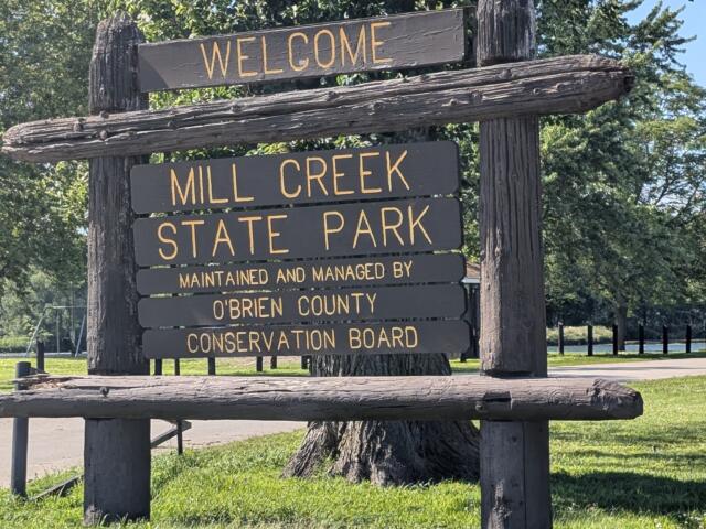 Wooden welcome sign for Mill Creek State Park with trees in the background. Maintained by O'Brien County Conservation Board.