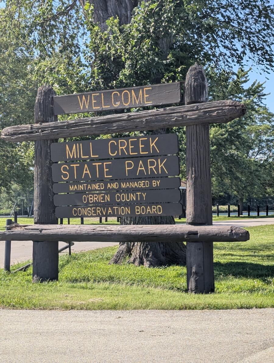 Wooden welcome sign for Mill Creek State Park with trees in the background. Maintained by O'Brien County Conservation Board.