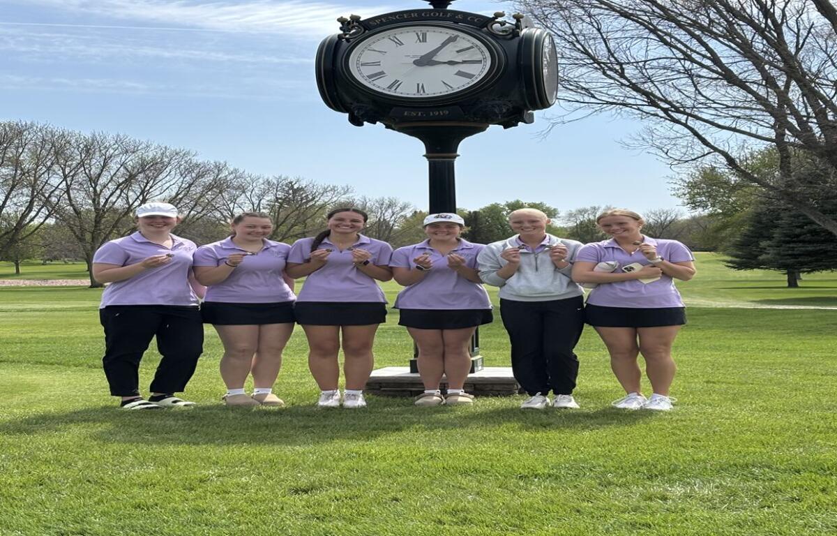 Six people in lavender polo shirts pose with medals in front of a large clock on a golf course.
