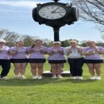 Six people in lavender polo shirts pose with medals in front of a large clock on a golf course.