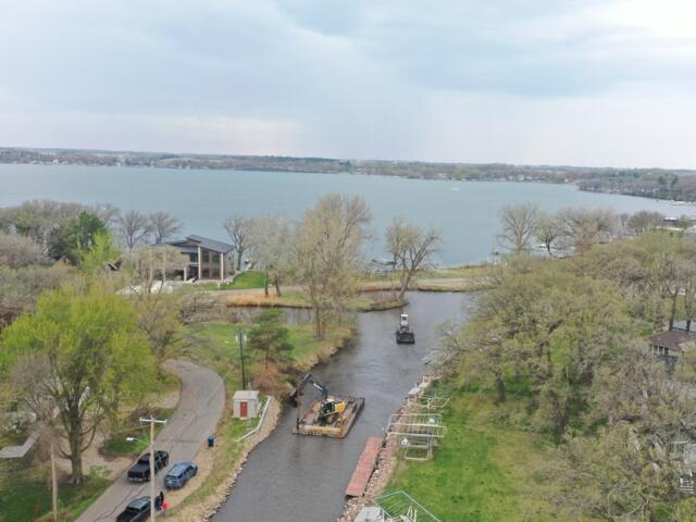 Aerial view of a river and canal with construction equipment on a barge in the water, trees lining the banks, and houses nearby.