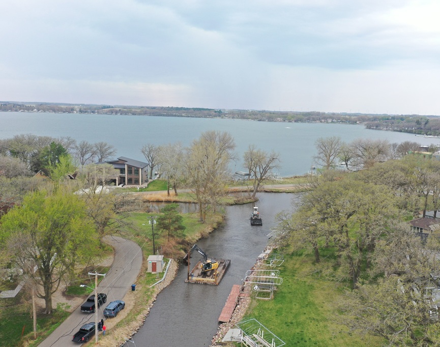 Aerial view of a river and canal with construction equipment on a barge in the water, trees lining the banks, and houses nearby.