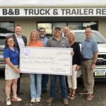 Eight people stand in front of a B&B Truck & Trailer Repair storefront, holding a large ceremonial check for $25,000 dated June 17, 2026.