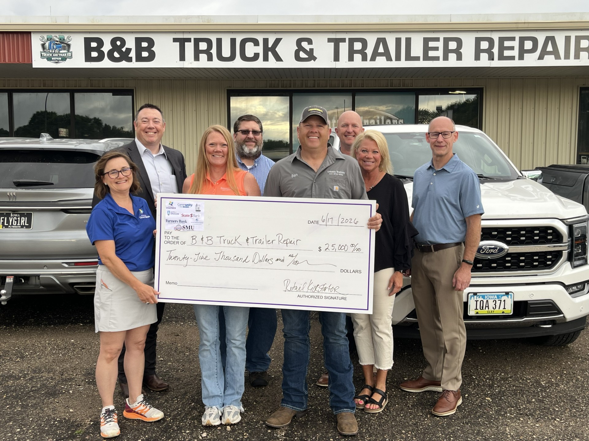 Eight people stand in front of a B&B Truck & Trailer Repair storefront, holding a large ceremonial check for $25,000 dated June 17, 2026.