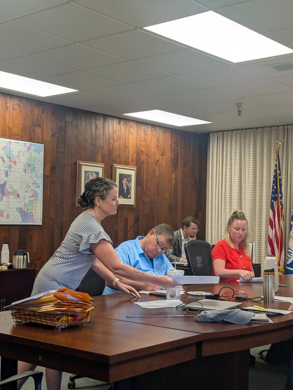 Woman standing and handing documents to a man at a large conference table during a formal meeting in a wood-paneled room with maps and portraits on the wall, and an American flag in the background.