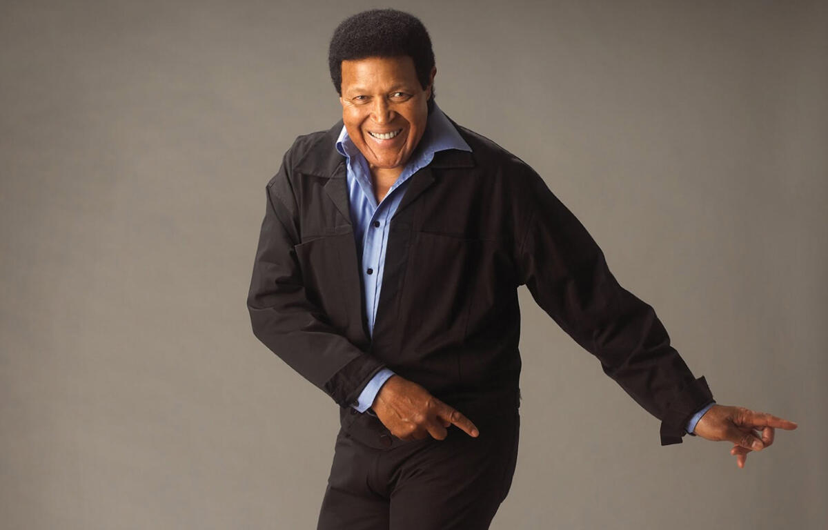 Smiling man in a black jacket and blue shirt points to the right against a neutral backdrop.