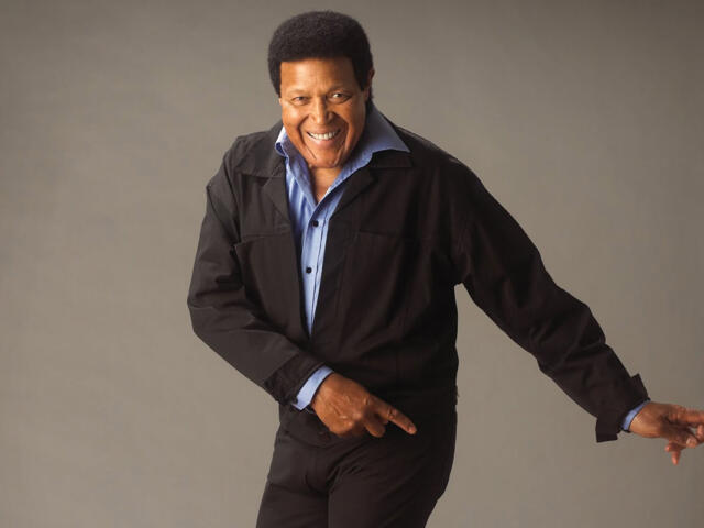 Smiling man in a black jacket and blue shirt points to the right against a neutral backdrop.