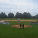 Baseball field scene with two teams lined up facing each other along the baselines; white uniforms on the left and green uniforms on the right, with two umpires standing near home plate.