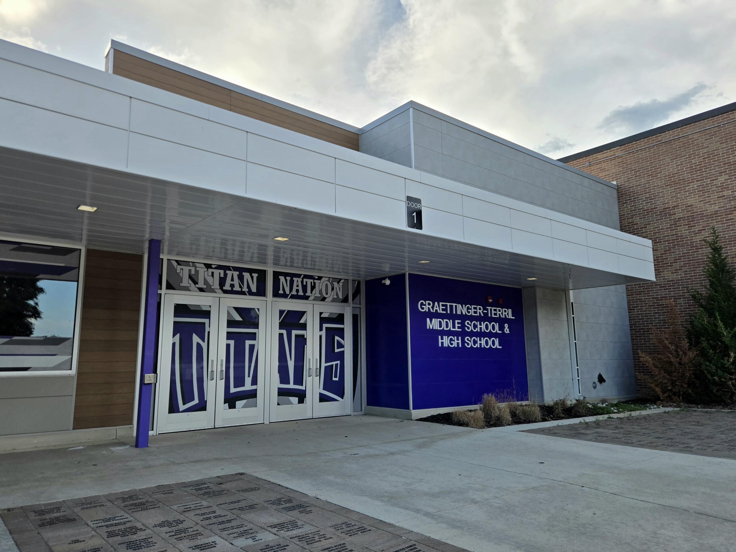 Front entrance of a modern school building with decorative glass doors featuring a large 'Titan Nation' graphic and a blue wall with the Graettinger–Terril Middle School & High School sign to the right.
