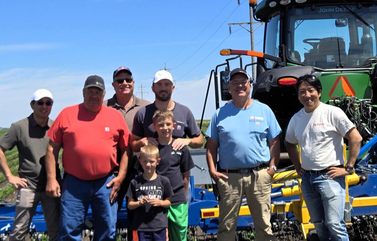 Group of men and two kids posing outdoors in front of a large green tractor and farming equipment under a blue sky.
