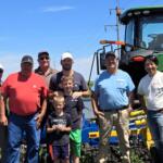 Group of men and two kids posing outdoors in front of a large green tractor and farming equipment under a blue sky.