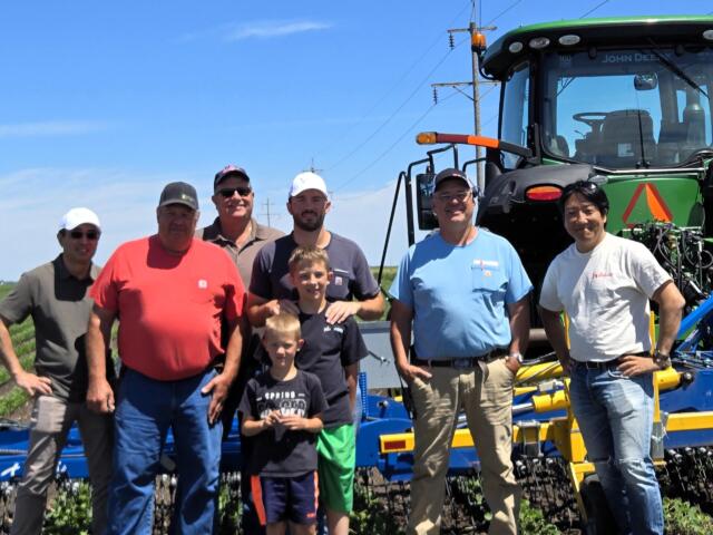 Group of men and two kids posing outdoors in front of a large green tractor and farming equipment under a blue sky.