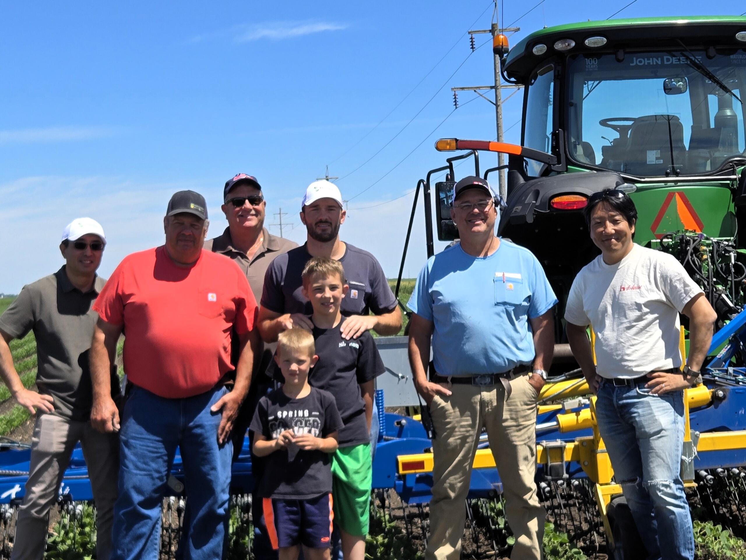 Group of men and two kids posing outdoors in front of a large green tractor and farming equipment under a blue sky.
