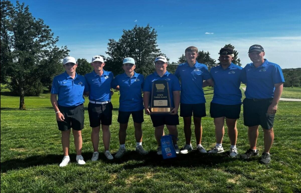 Seven male golfers in matching blue polo shirts and shorts stand on a grassy golf course, holding a trophy that reads 'State Third Place' amid a sunny backdrop with trees behind them.