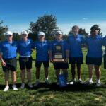 Seven male golfers in matching blue polo shirts and shorts stand on a grassy golf course, holding a trophy that reads 'State Third Place' amid a sunny backdrop with trees behind them.