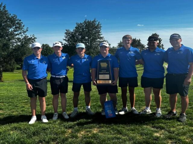Seven male golfers in matching blue polo shirts and shorts stand on a grassy golf course, holding a trophy that reads 'State Third Place' amid a sunny backdrop with trees behind them.