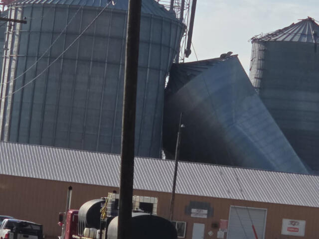 Tilted metal grain silo leaning against neighboring silos at an industrial facility, with a brown warehouse in the foreground and utility poles nearby.
