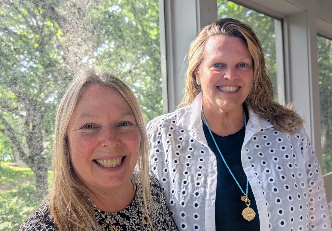 Two women smiling at the camera indoors beside large windows with greenery outside. for context: casual portrait of colleagues.