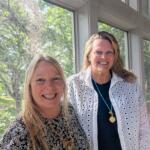 Two women smiling at the camera indoors beside large windows with greenery outside. for context: casual portrait of colleagues.