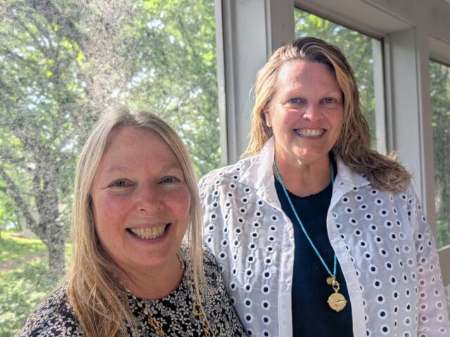 Two women smiling at the camera indoors beside large windows with greenery outside. for context: casual portrait of colleagues.