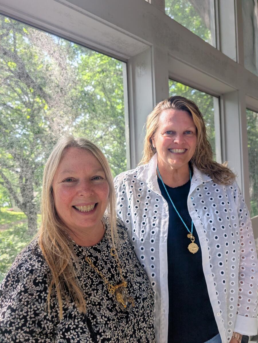 Two women smiling at the camera indoors beside large windows with greenery outside. for context: casual portrait of colleagues.
