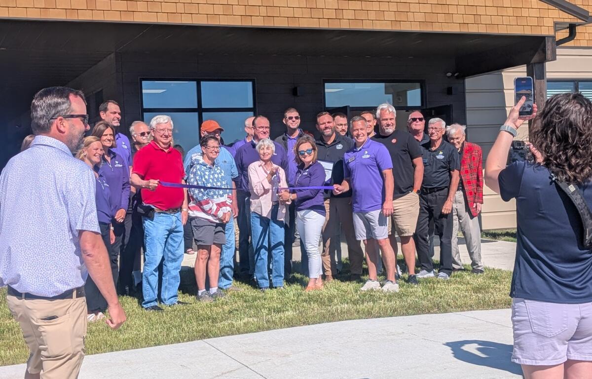 Group of people posing for a ribbon-cutting photo outside a tan-brick building during a grand opening.
