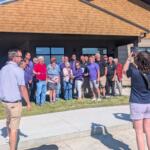Group of people posing for a ribbon-cutting photo outside a tan-brick building during a grand opening.