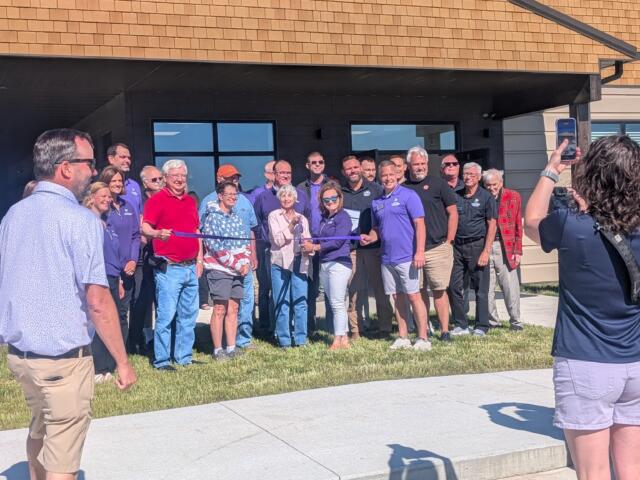 Group of people posing for a ribbon-cutting photo outside a tan-brick building during a grand opening.