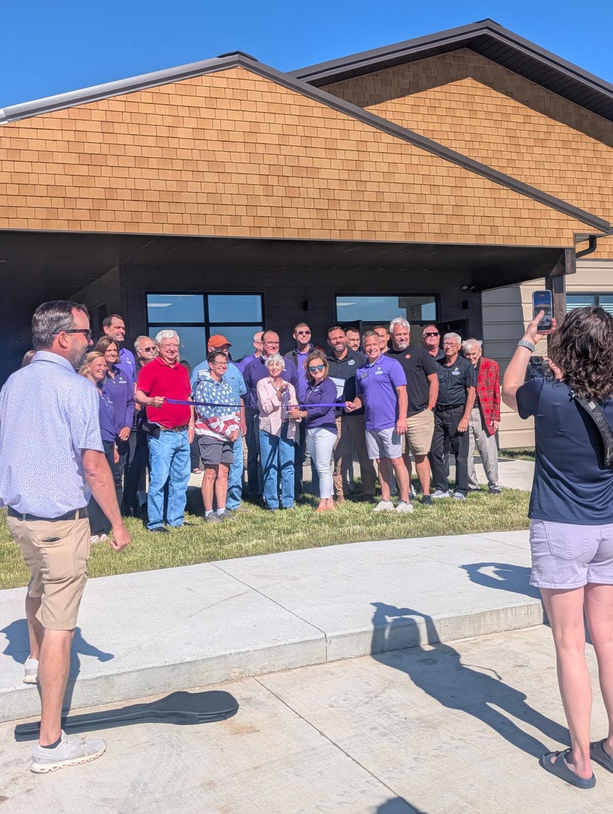 Group of people posing for a ribbon-cutting photo outside a tan-brick building during a grand opening.