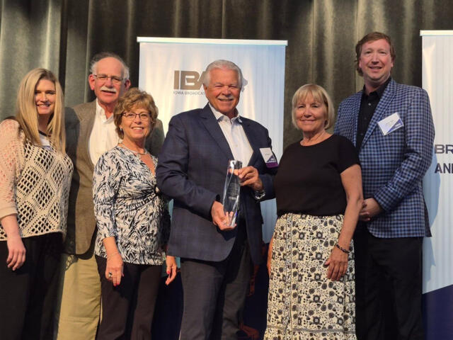 Group of seven people posing onstage at an awards event, one man in a suit holds a glass trophy, smiling for the photo.