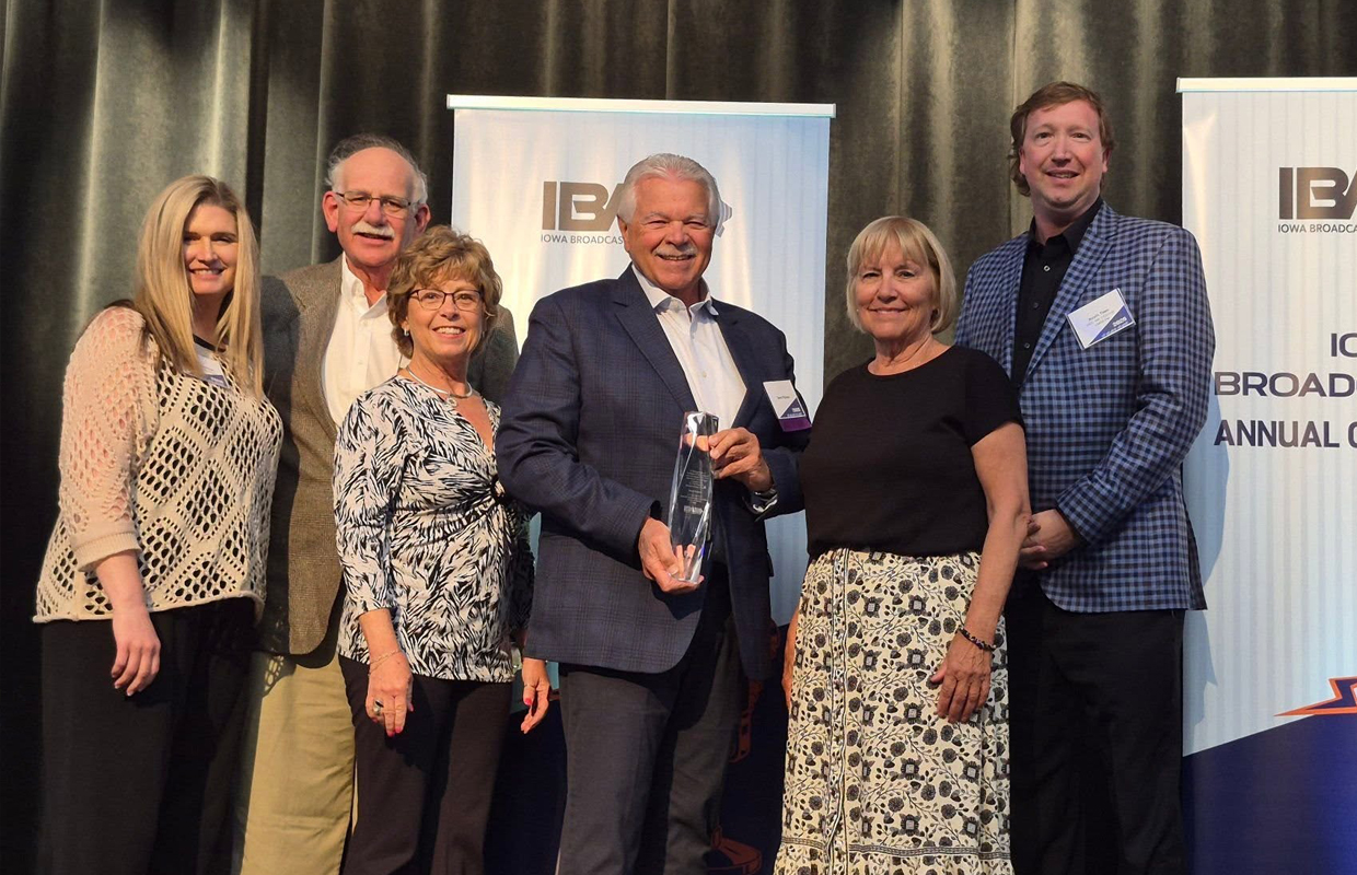 Group of seven people posing onstage at an awards event, one man in a suit holds a glass trophy, smiling for the photo.