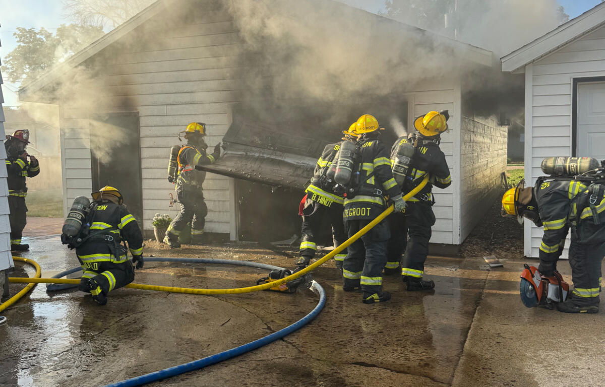 Firefighters in full gear work to extinguish a house garage fire, yellow hoses on a wet driveway as smoke billows from the structure behind a collapsed door frame.